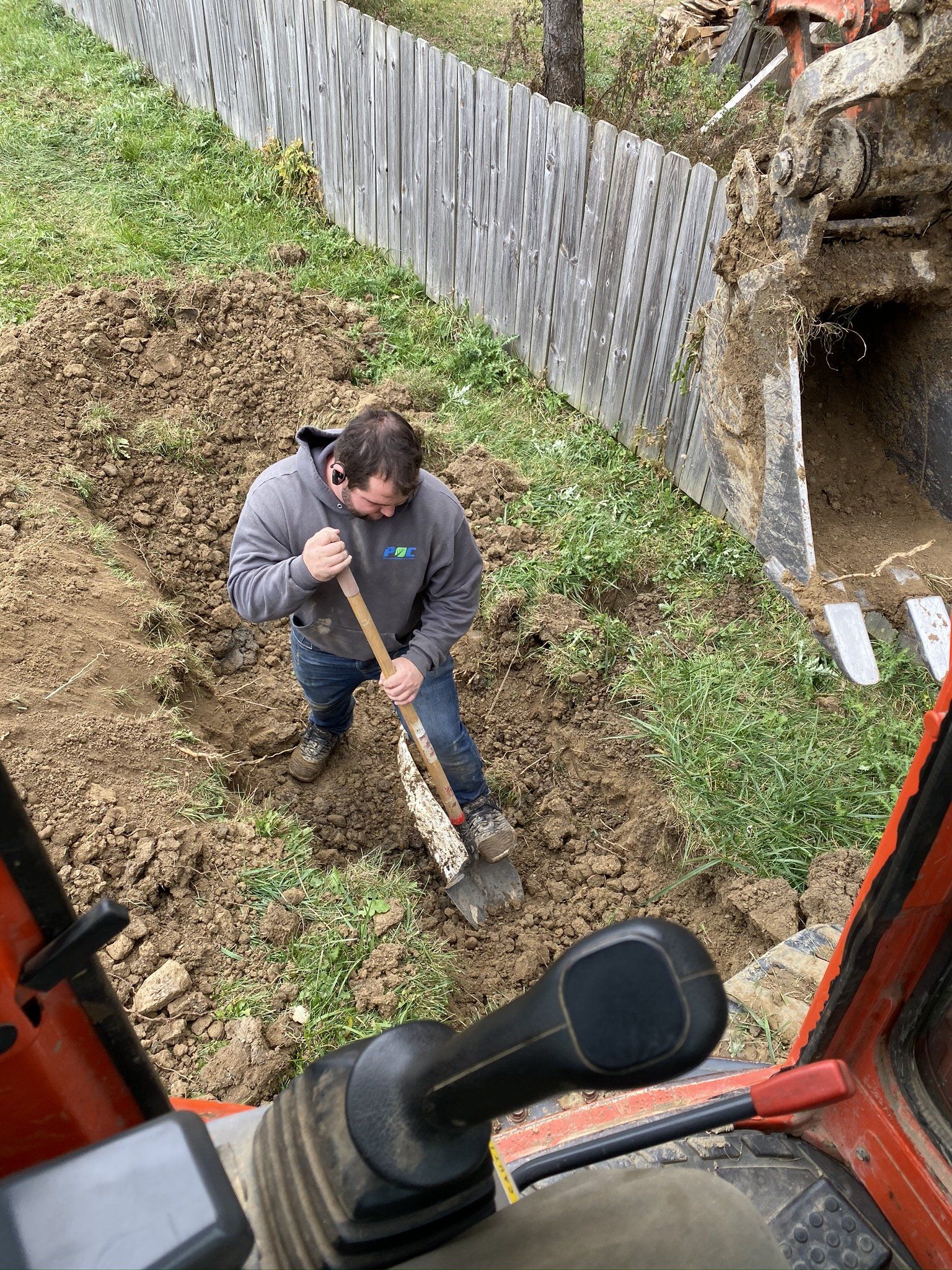 Man shovels dirt beside a fence and an excavator; outdoor, daytime.