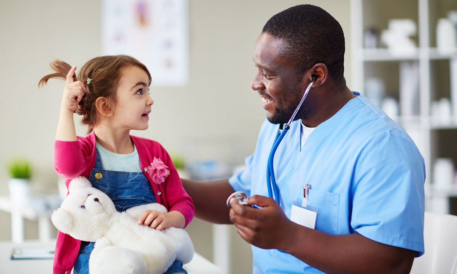 A doctor with a stethoscope examines a child holding a teddy bear in a clinic.