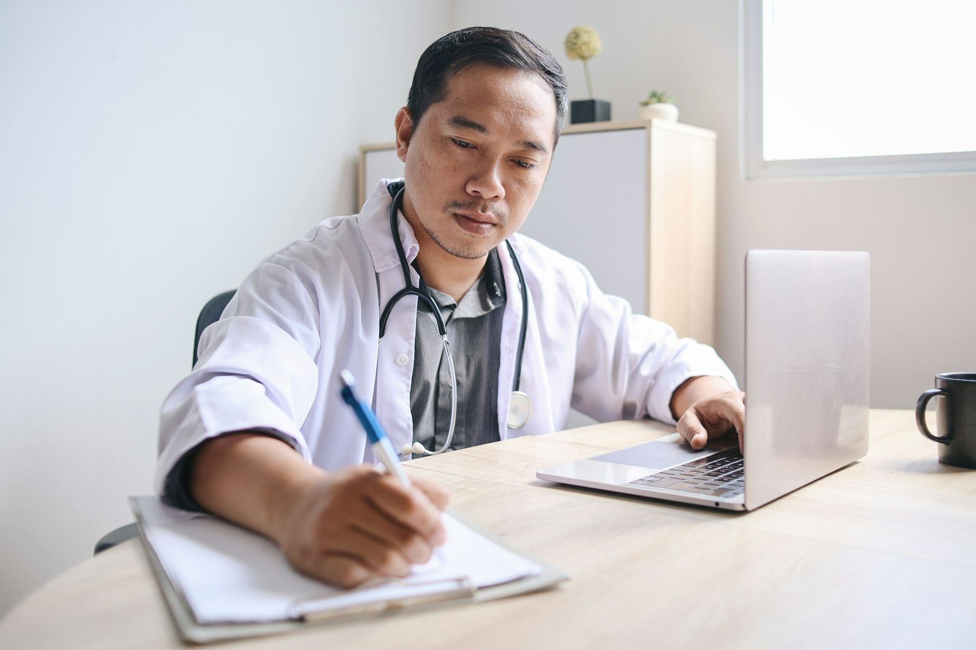 Doctor in white coat, stethoscope, writing on notepad, using laptop at desk.