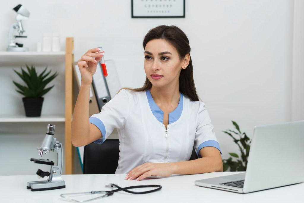 Doctor examines vial in a medical office, with microscope and laptop.