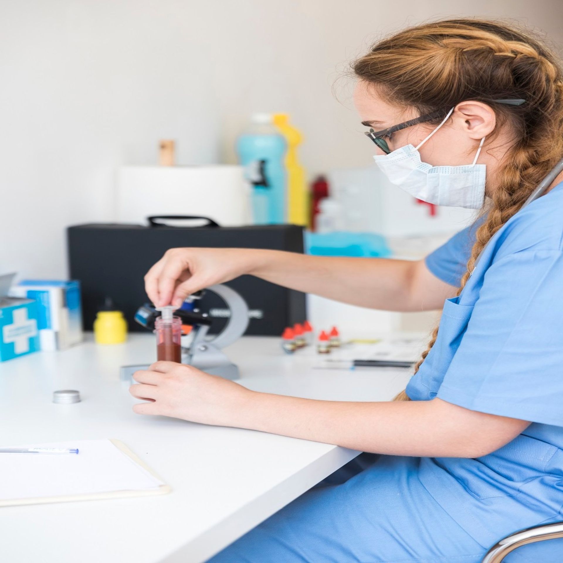 Lab worker in blue scrubs, mask, and glasses, examining a vial of blood with a microscope.