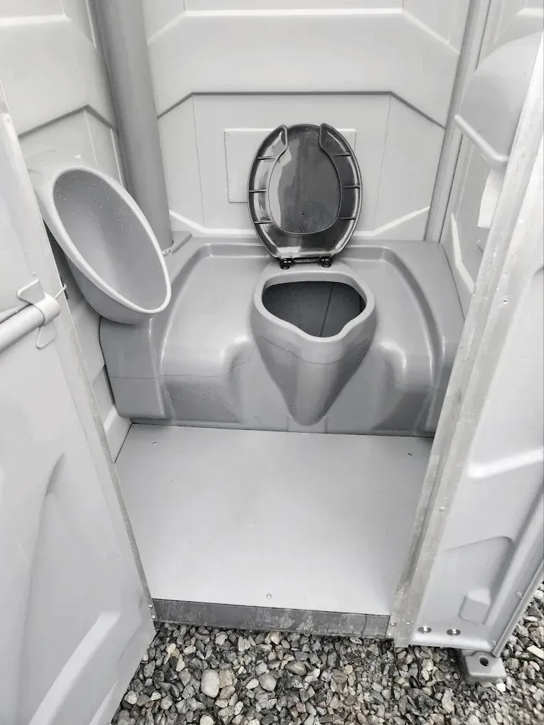 Portable toilet interior with open seat and urinal, viewed from the doorway, gravel outside.