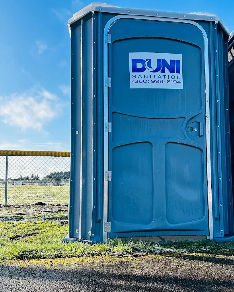 Blue DUNI portable toilet beside a fence on a grassy field under a clear sky
