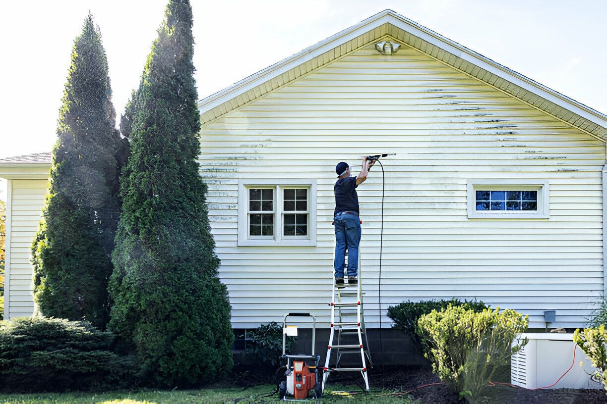 Man on ladder washing siding of a white house with power washer.