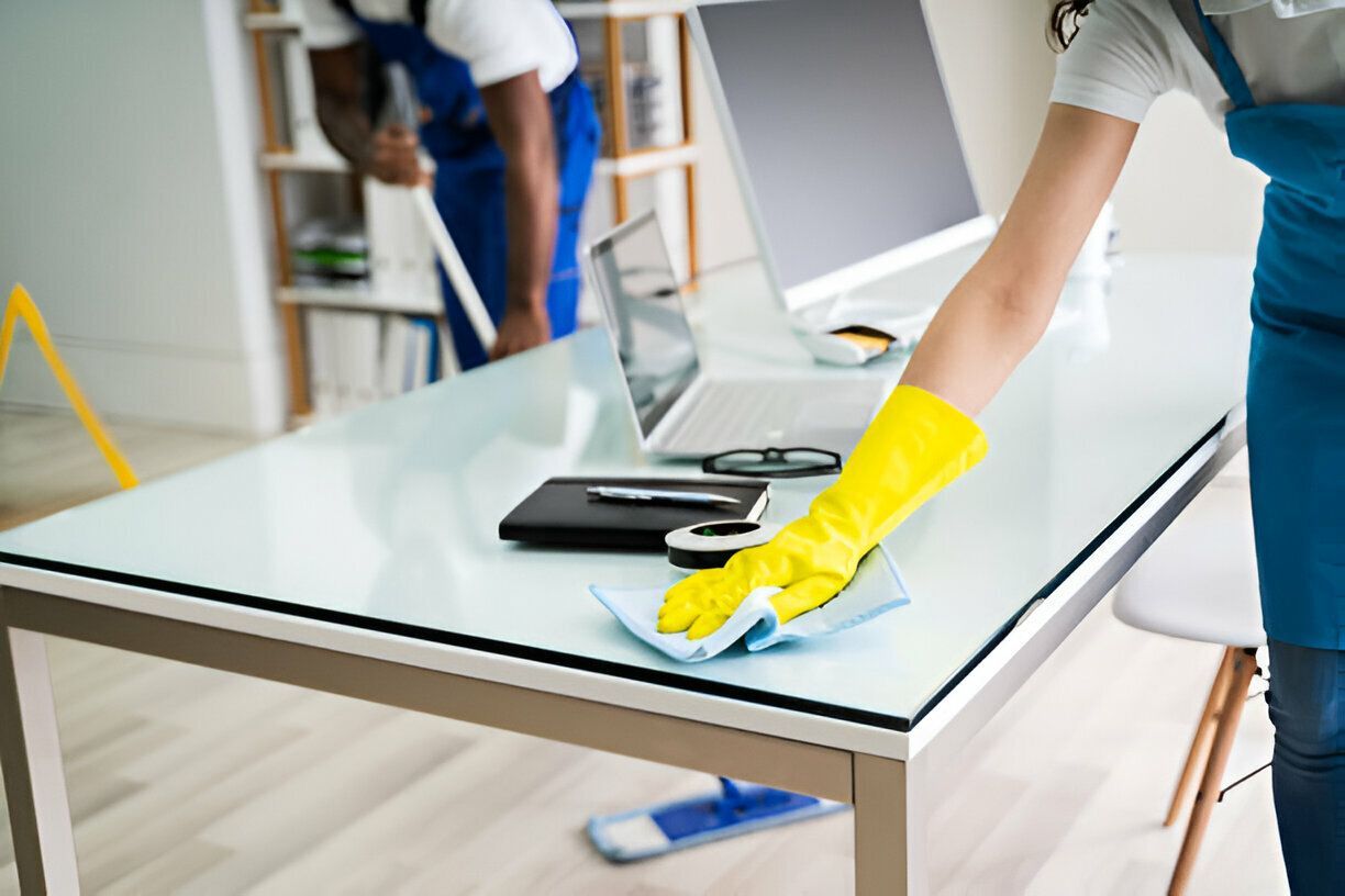 Person wearing yellow gloves wiping a white office desk; another person cleans in the background.