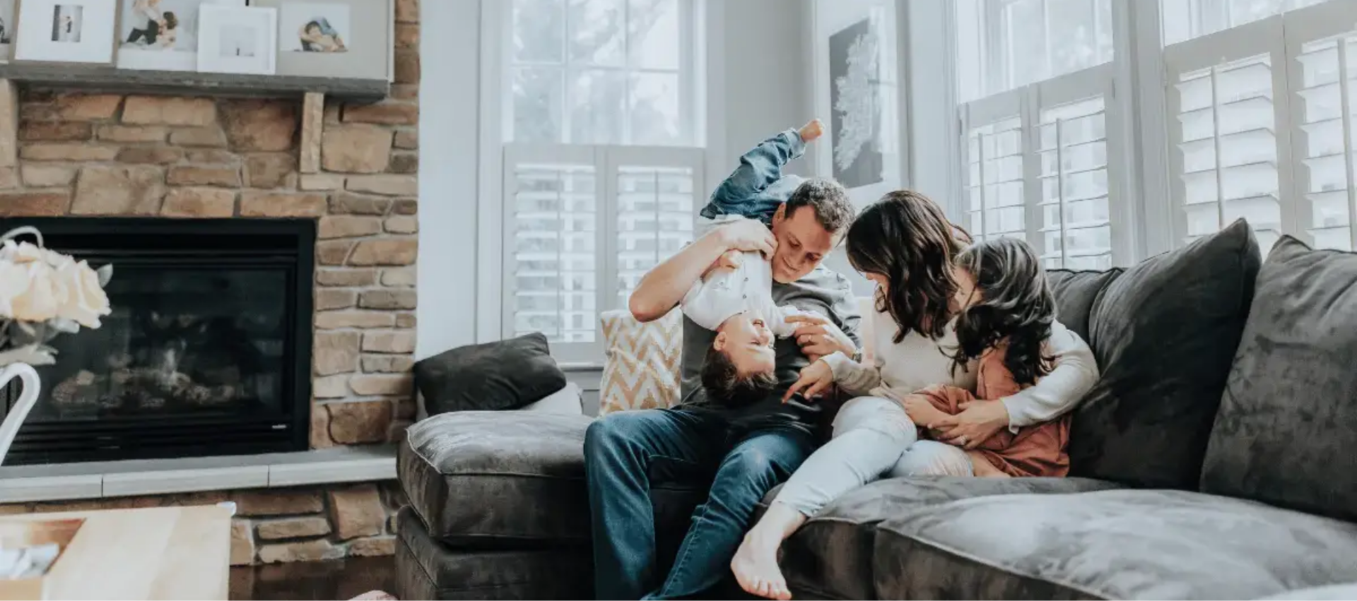 Family sitting on a couch by a fireplace, embracing and looking at a baby in a cozy living room.