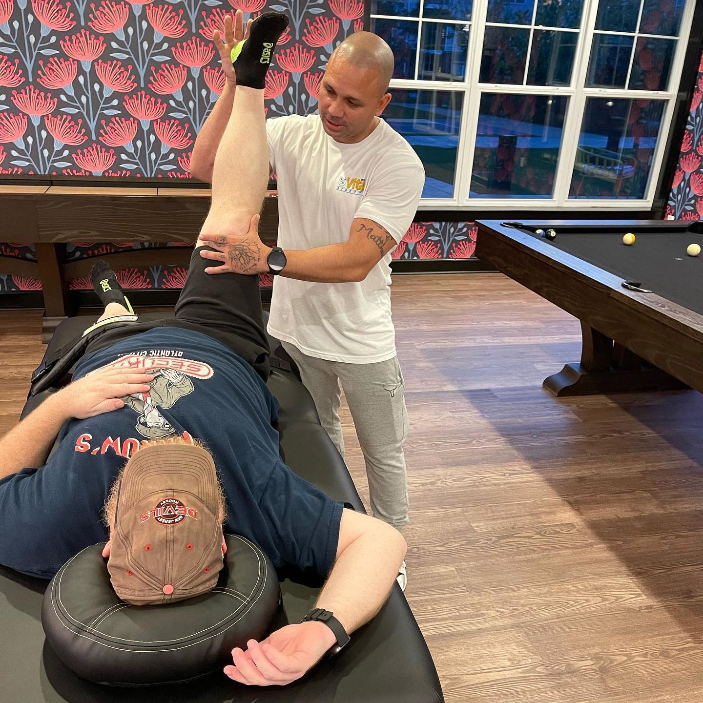 A man is stretching another man 's leg in a room with a pool table.