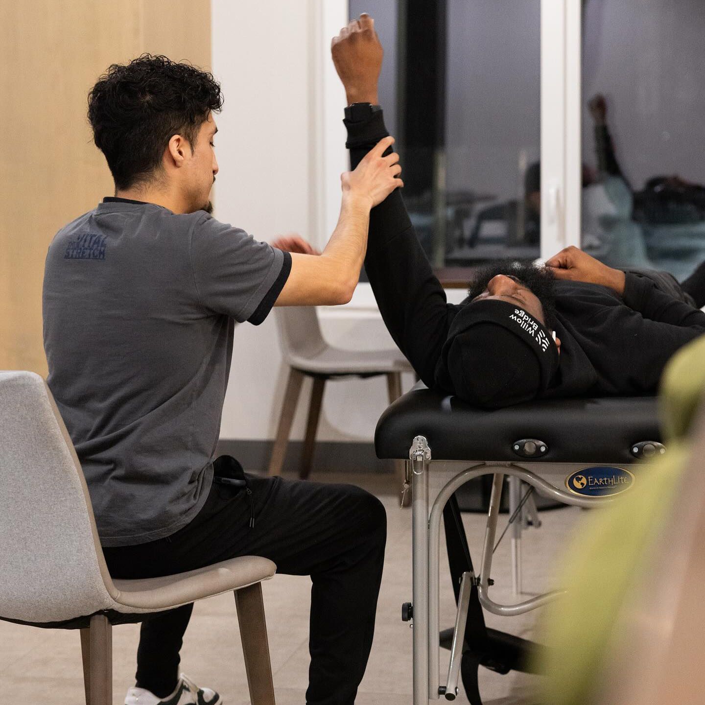 A man is sitting in a chair helping another man stretch his arm