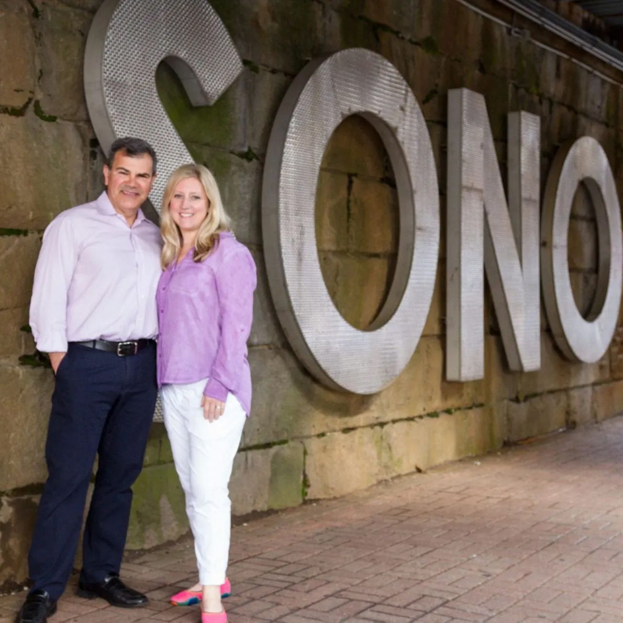 A man and a woman are standing in front of a large sign that says sono.