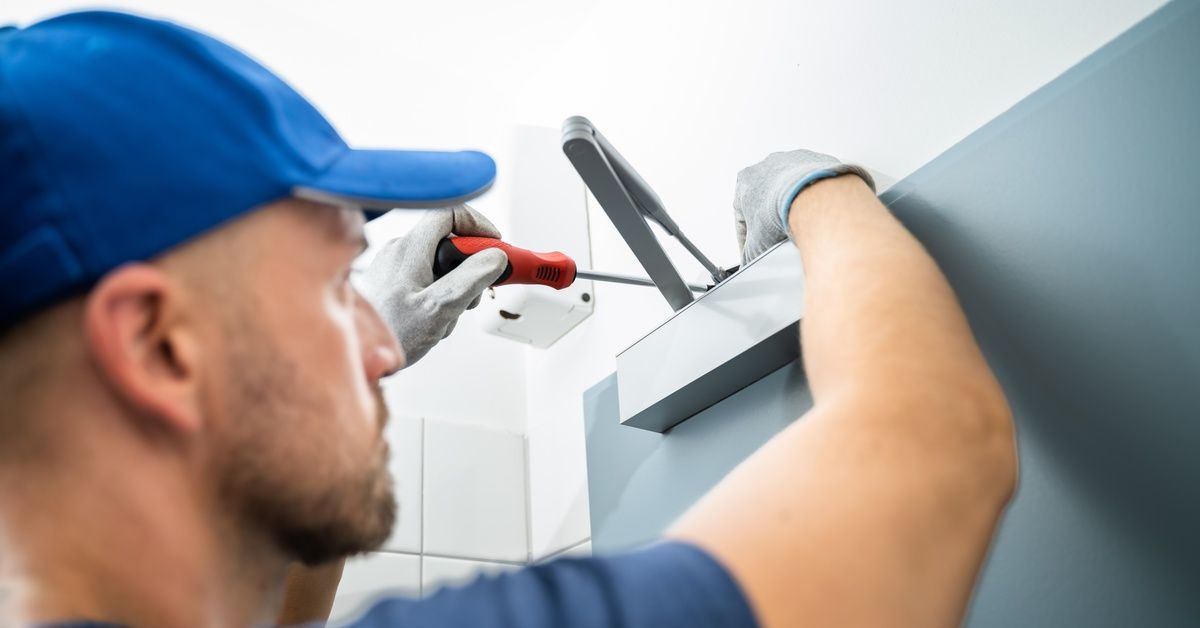 A repair man in white gloves and a blue hat using a screwdriver to repair an automatic door.