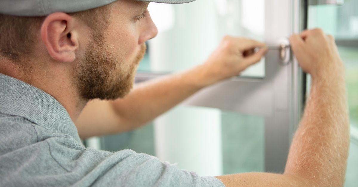 A worker wearing a gray shirt and hat uses tools to repair an automatic sliding glass door.