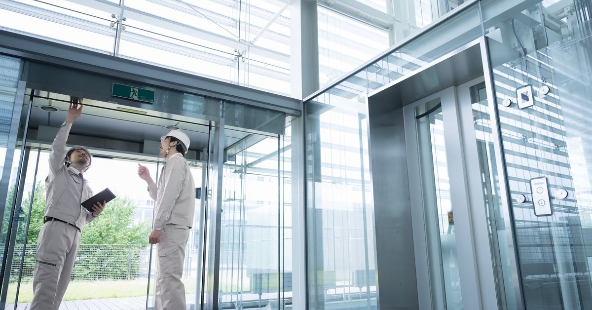 Two men wearing white uniforms and hard hats inspecting an automatic door inside a modern glass buil