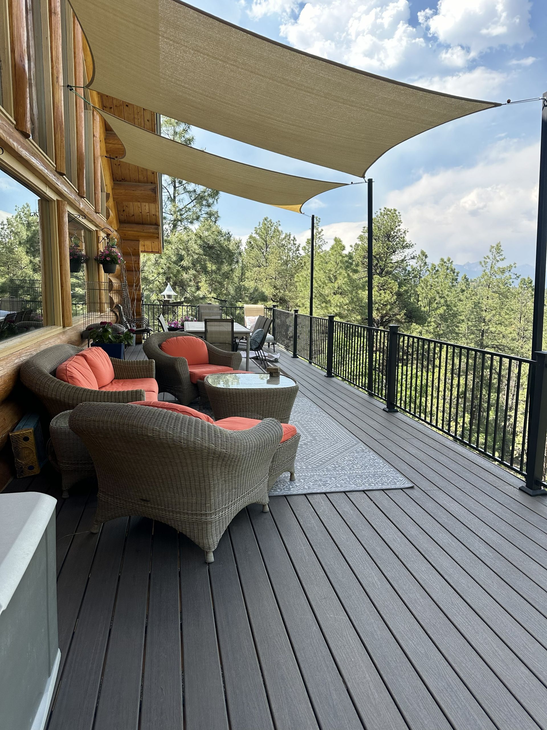 Outdoor deck with wicker furniture, orange cushions, and sun shade. View of trees and sky.