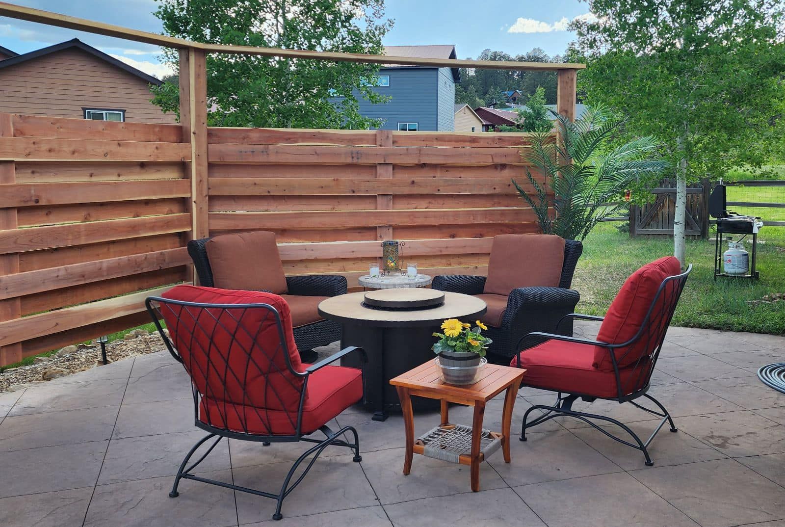 Patio with red chairs, a small table, and a round fire pit, bordered by a wooden fence.