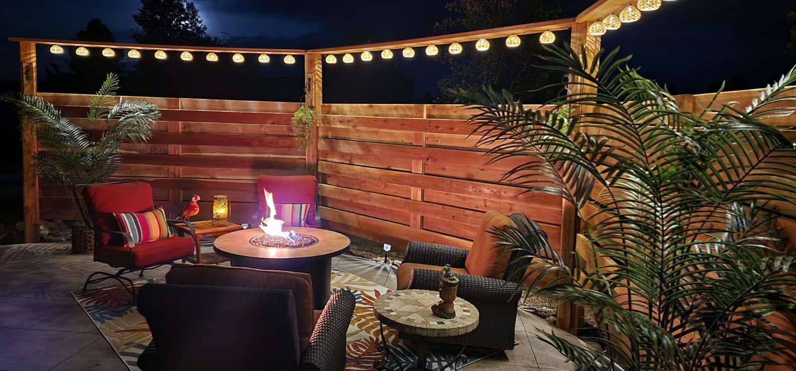 Cozy outdoor seating area at night with a fire pit, red chairs, string lights, and a wooden fence.