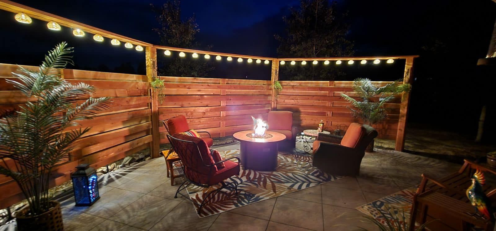 Outdoor patio at night with fire pit, seating, string lights, and wood fence.