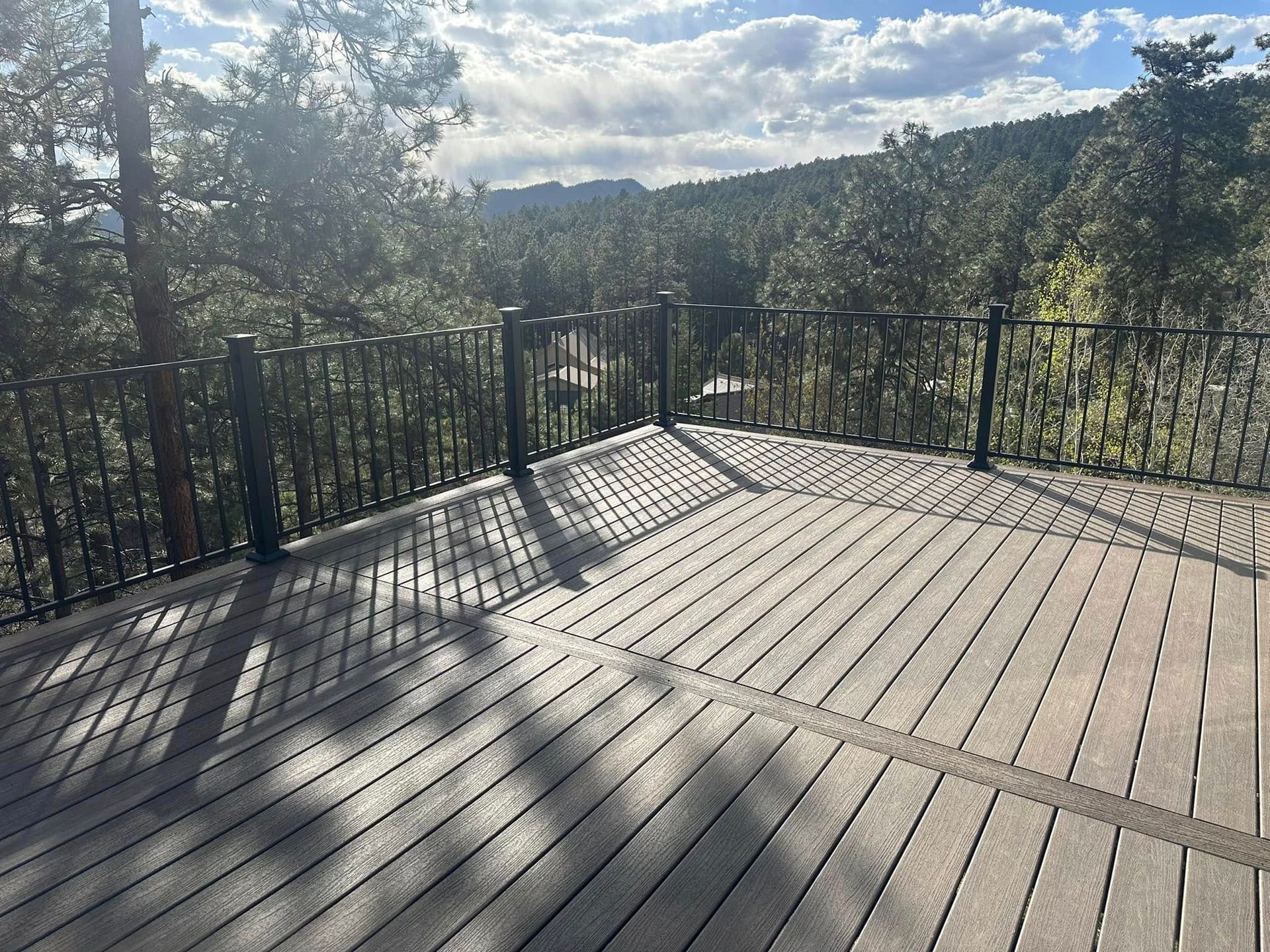 Wooden deck with black railing overlooking a forest, shadows cast by trees, sunny day.