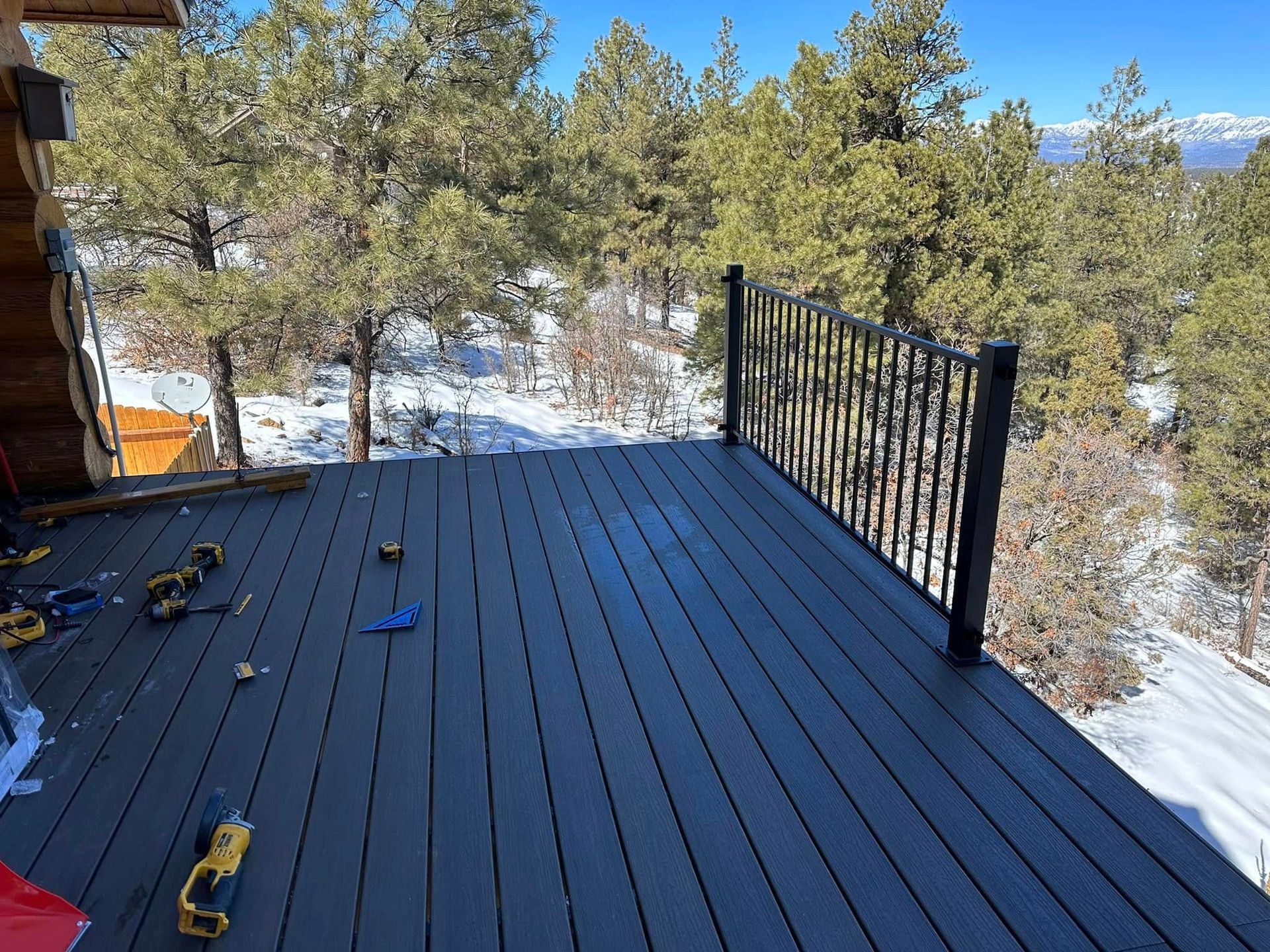 Black composite deck with black railing in a snowy forest setting on a sunny day.