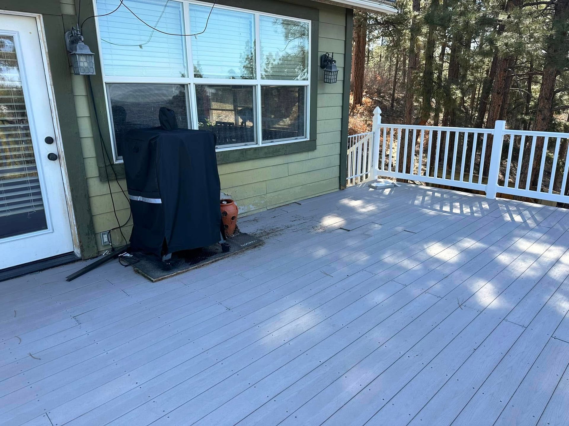 Grey wooden deck with white railing. A covered grill stands near the house. Sunny outdoors.