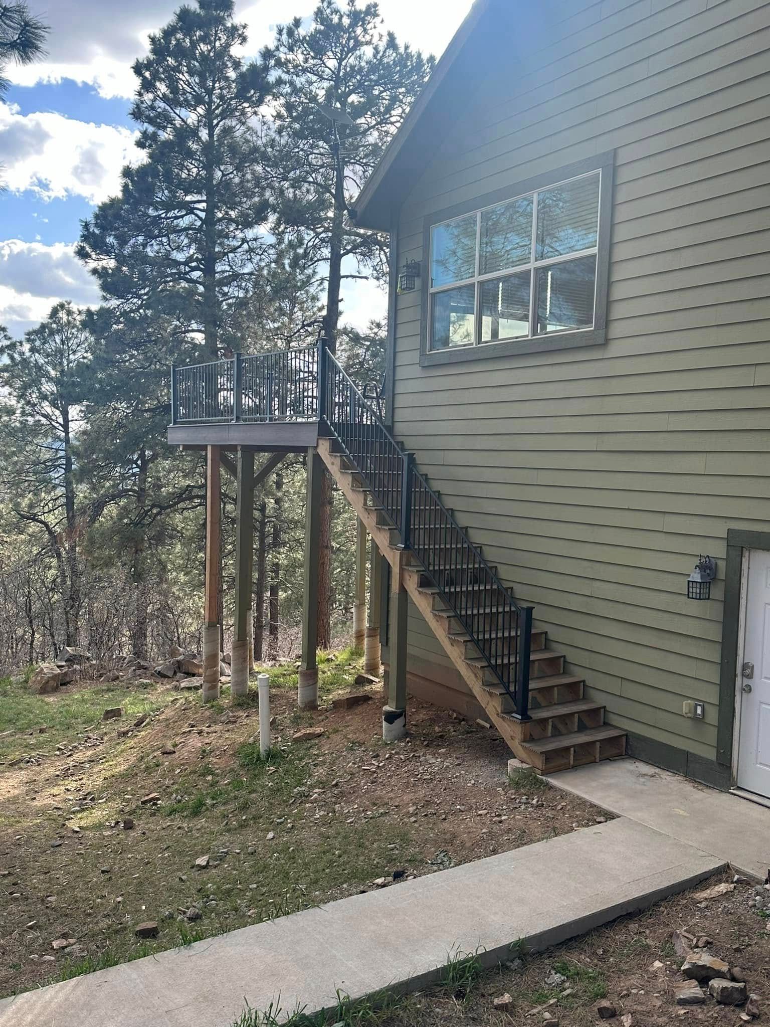 Exterior of a house with stairs leading to a deck raised on poles. Light green siding, dark railing, concrete path.