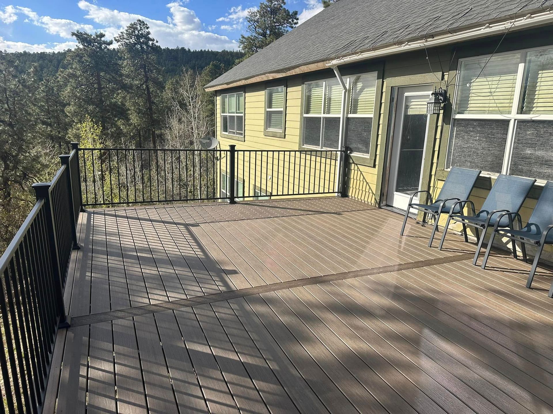 Wooden deck with black railing attached to a yellow house, chairs sit at the door.