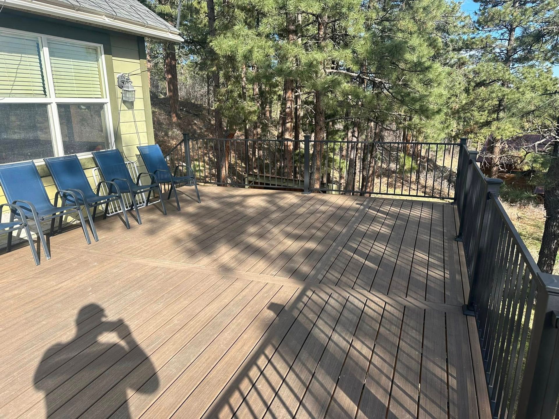 Wooden deck with blue chairs, surrounded by trees and railing. Shadow of person taking photo.