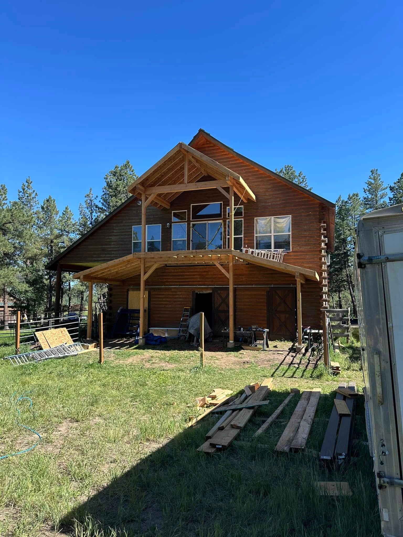 Two-story log home under construction with wooden beams, surrounded by trees and grass under a blue sky.