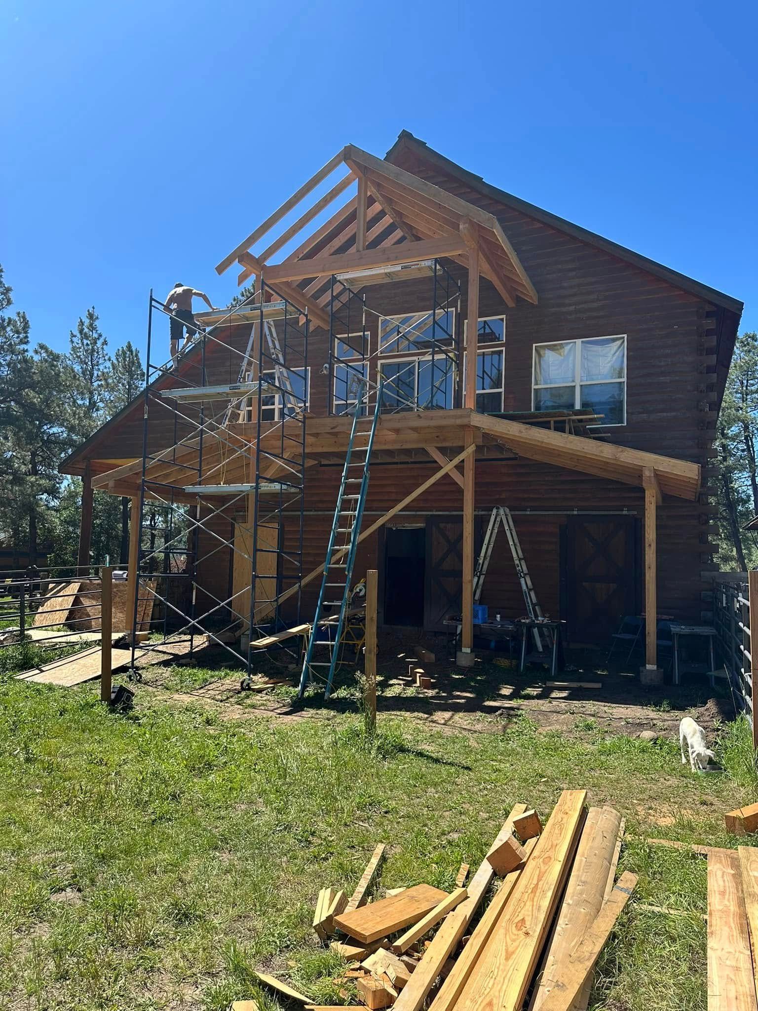 Construction on a brown log cabin home with workers, ladders, and lumber on a sunny day.