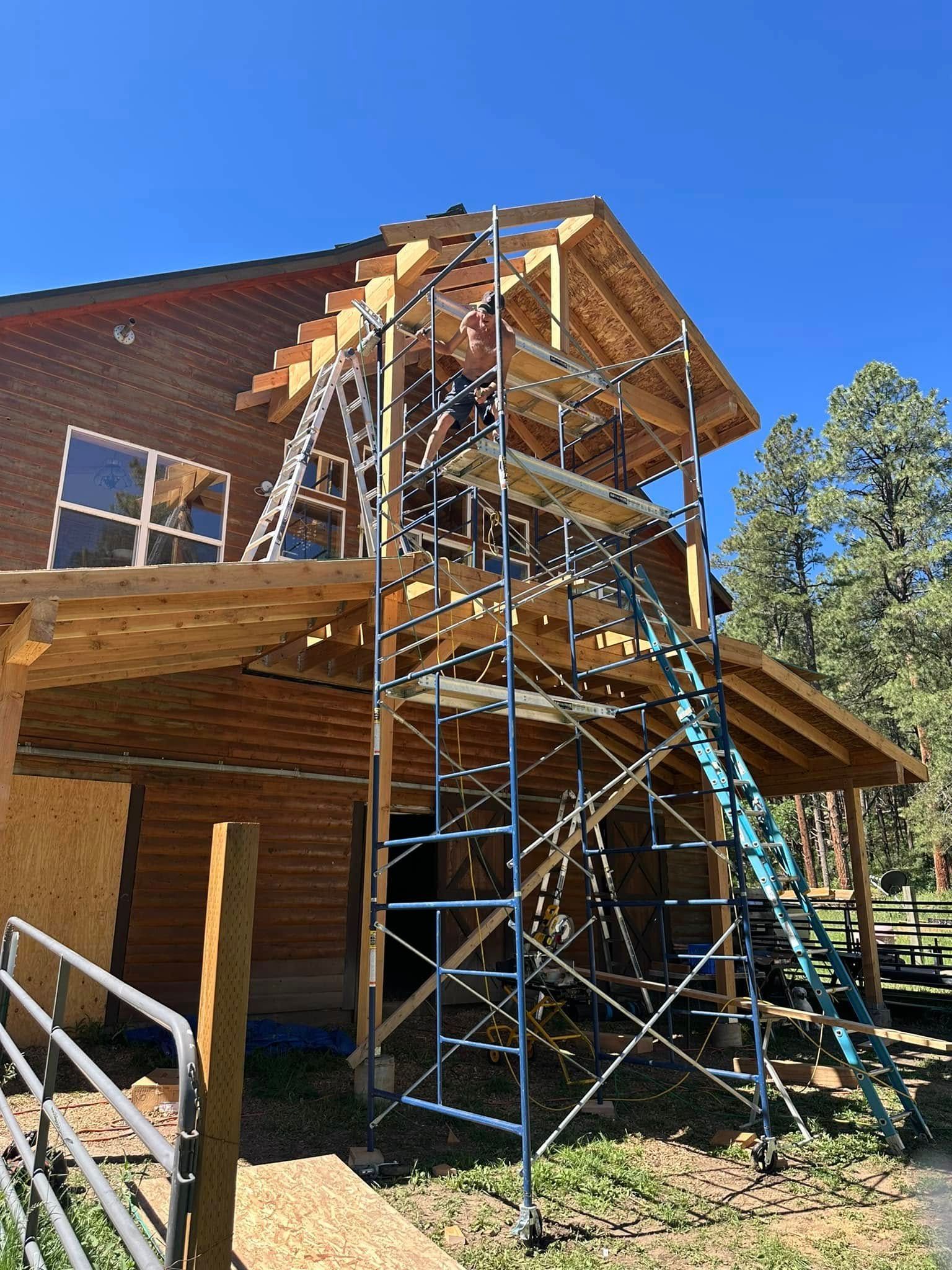 Construction scaffolding at a two-story house with a partially built roof, surrounded by trees.