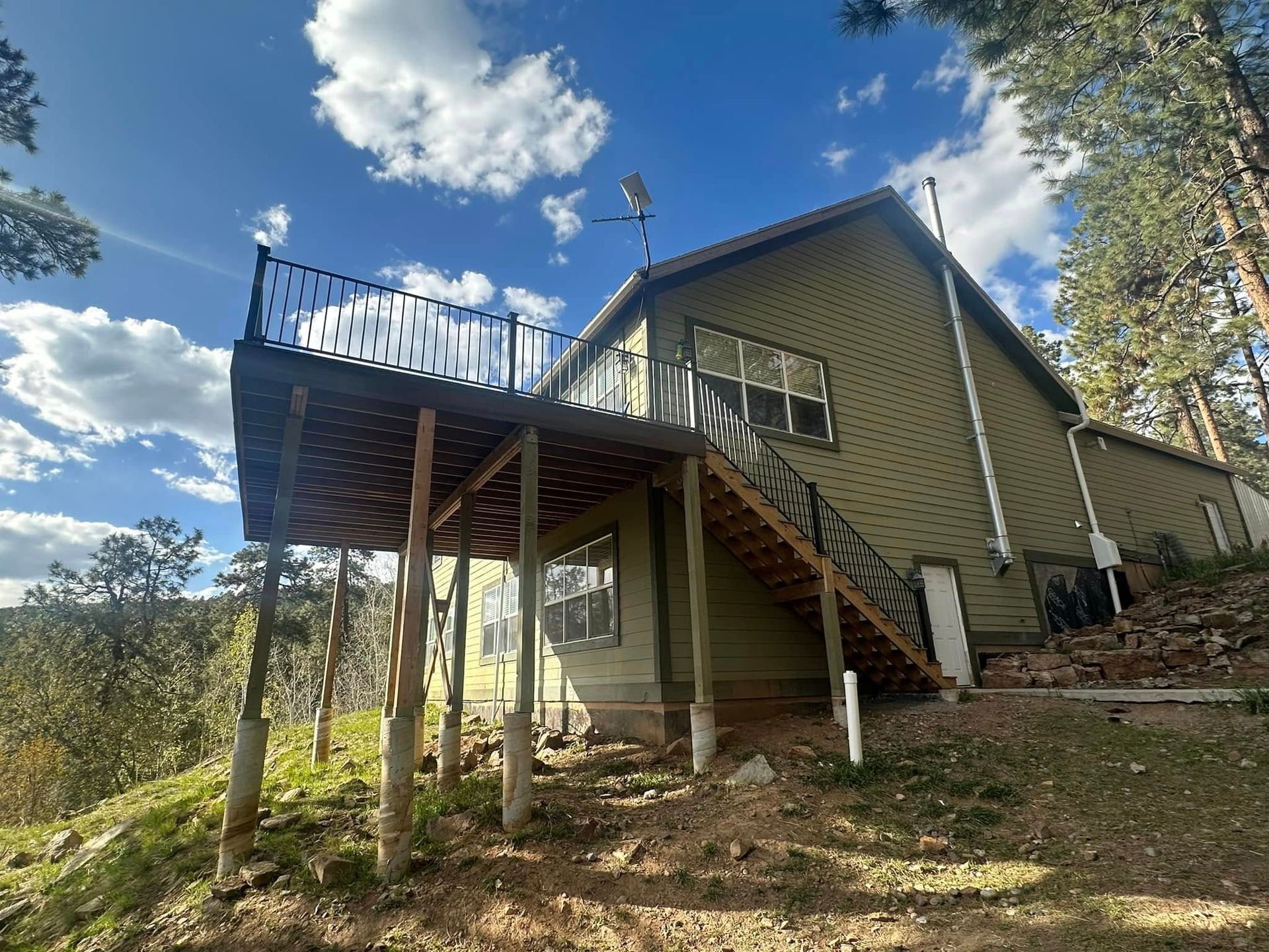 Green house with deck, stairs, and railing. Set on a hill, with a blue sky and some trees.
