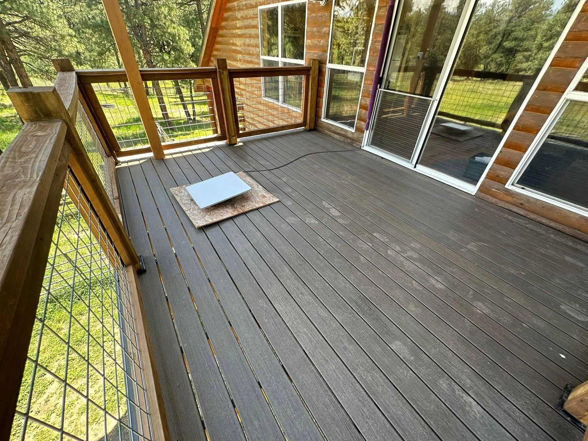 Wooden deck with dark gray planks, surrounded by a railing, with trees visible in the background.