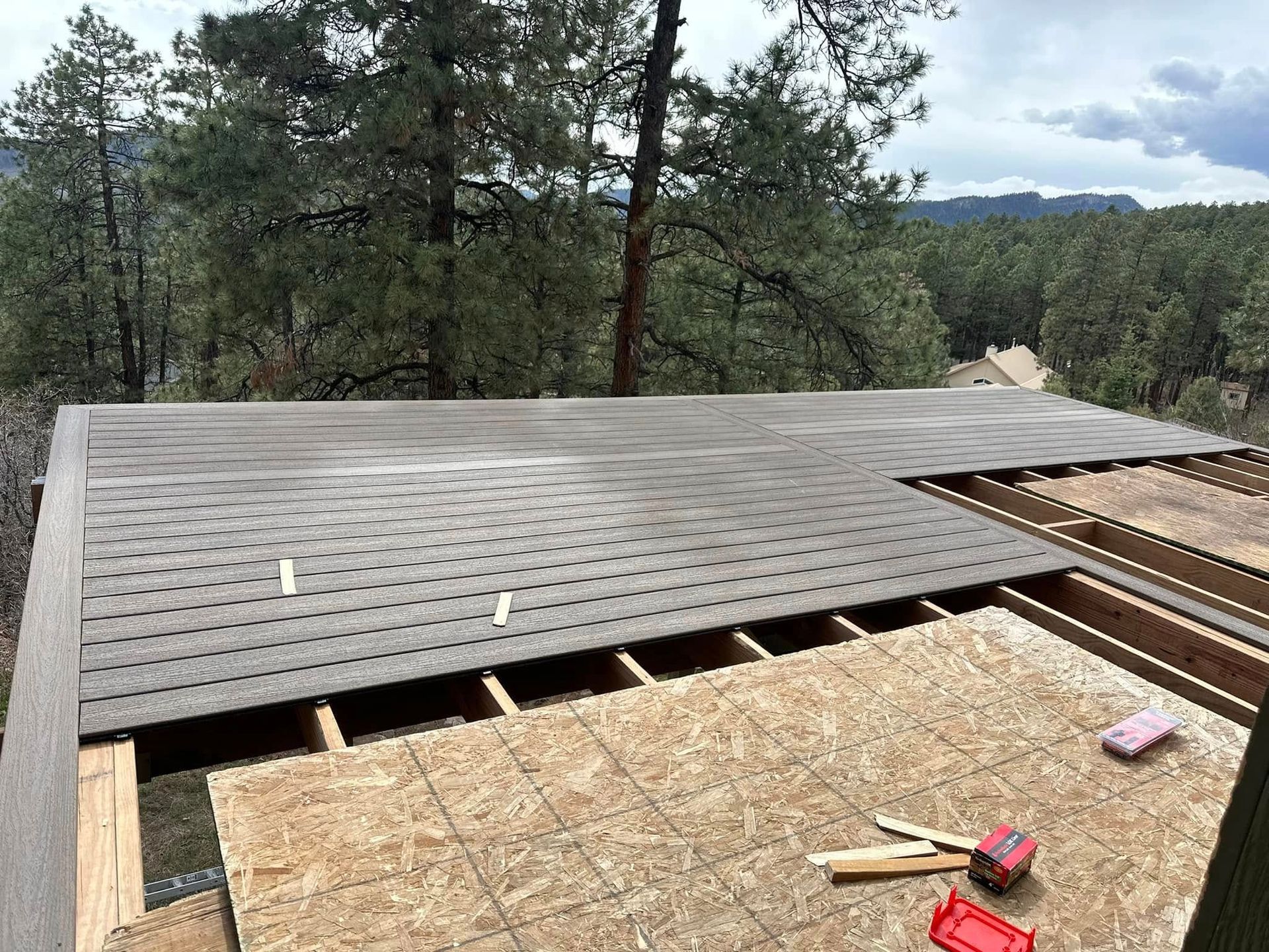 Rooftop construction with wood framing and shingle installation against a forest backdrop.