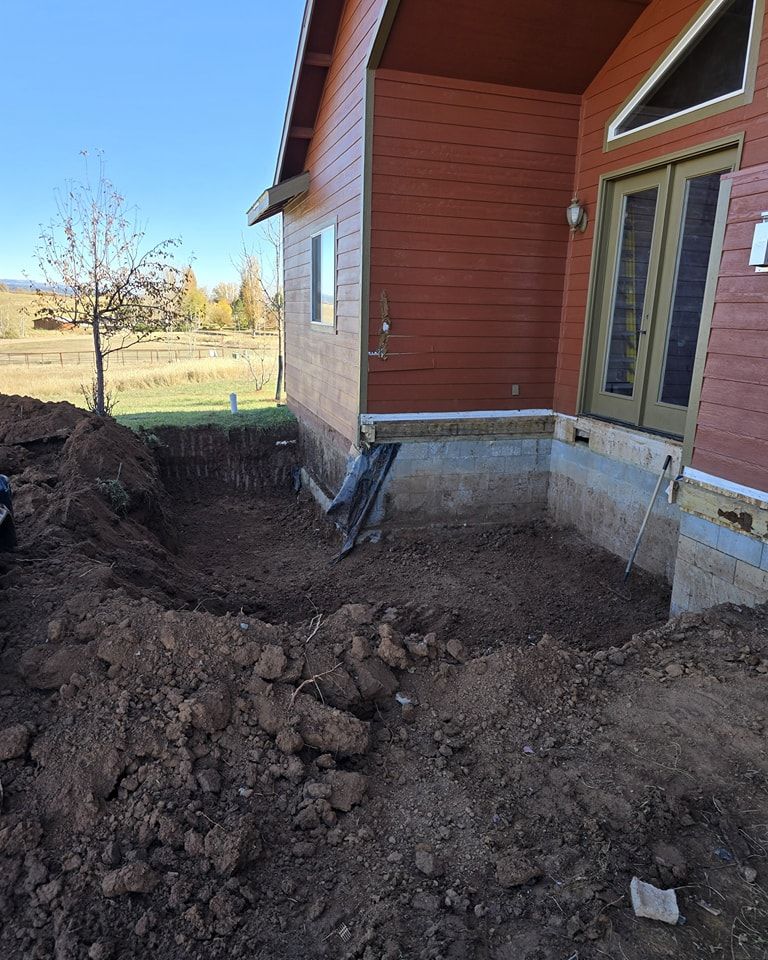 Excavation next to a reddish house. Dirt pile, exposed foundation, and French doors. Clear sky.