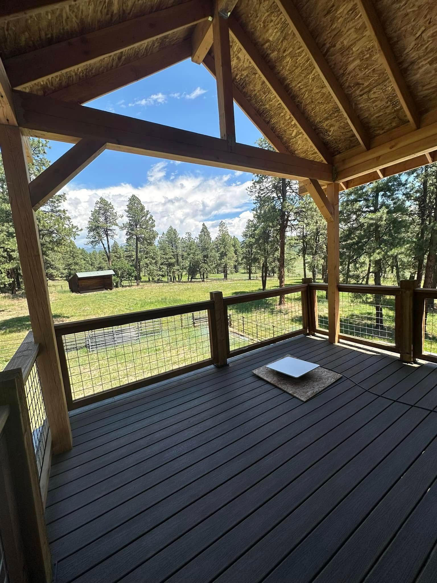 Wooden deck overlooking a green field and forest, under a covered structure with blue sky.