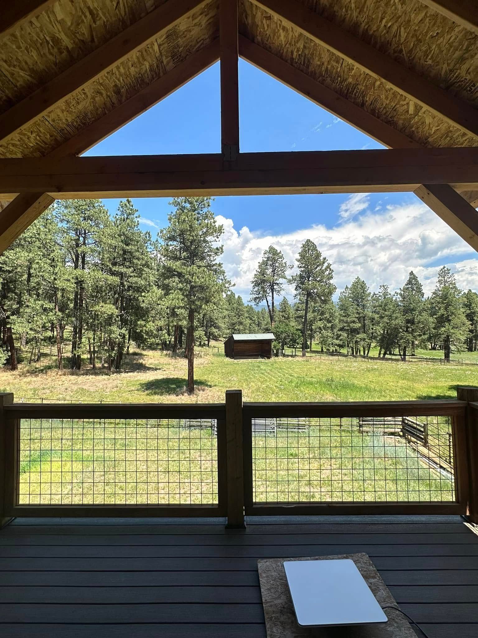 Wooden porch with view of a grassy field, trees, and a small cabin, blue sky peeking through.