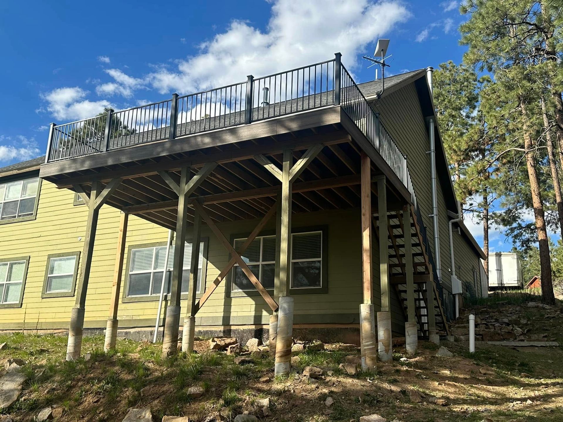 Wooden deck on a two-story building with a railing, supported by posts on a hillside, blue sky.