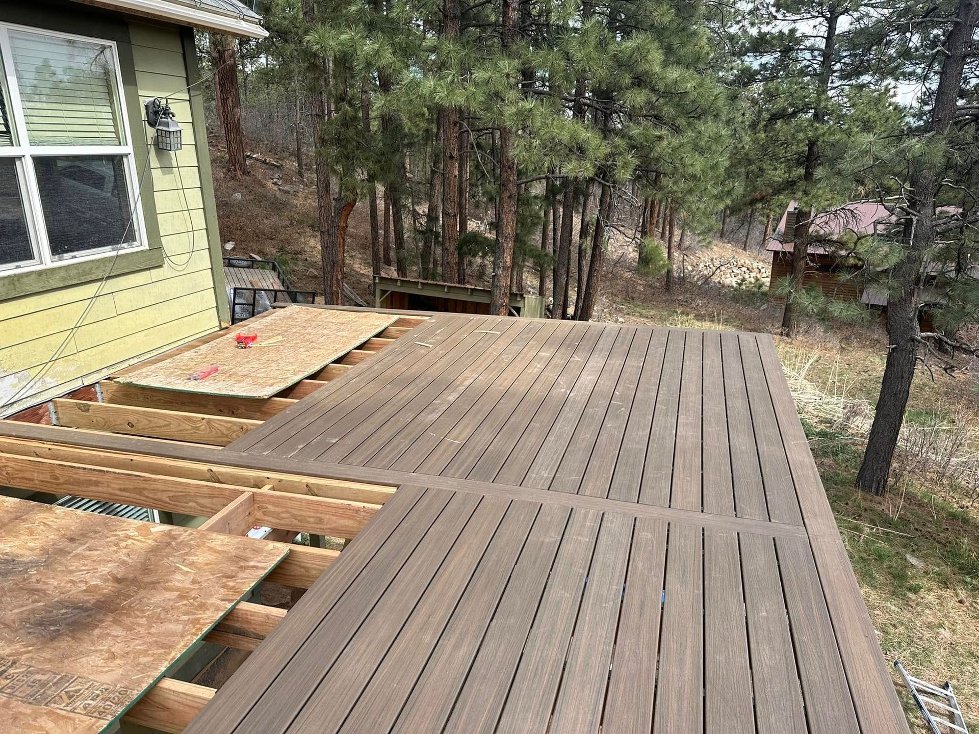 Deck being built on the side of a house, with composite boards installed, partially covered with plywood, and trees in the background.