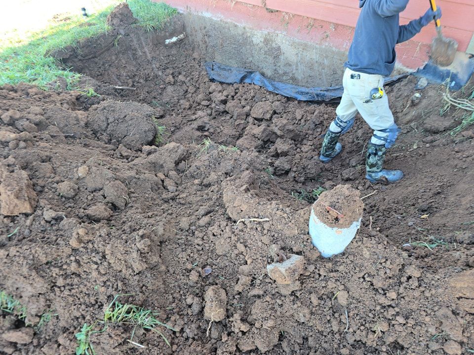 Person digging in dirt near a building foundation, using a shovel; muddy ground.