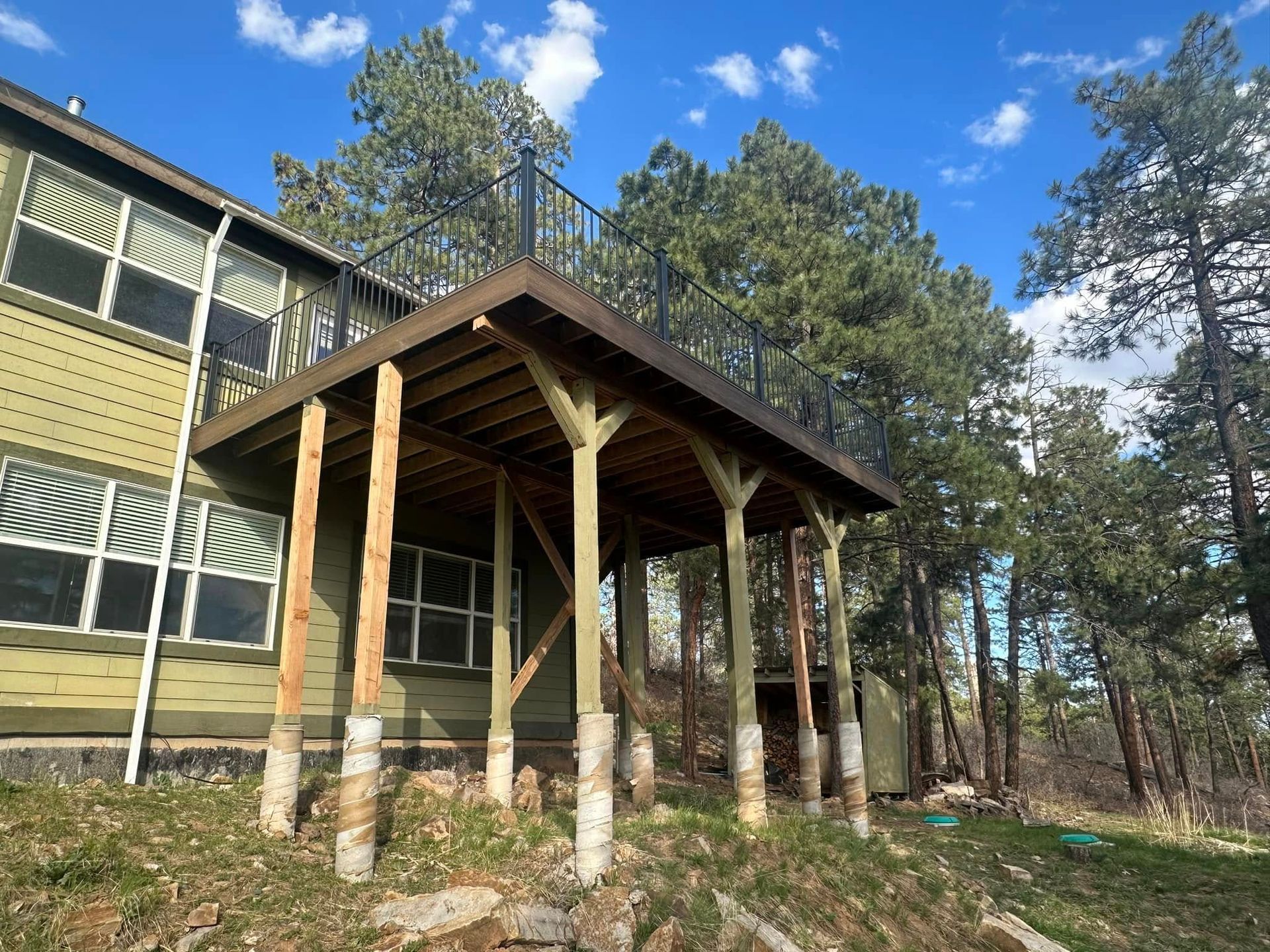 A covered wooden deck attached to a green house, supported by pillars, set in a wooded area.