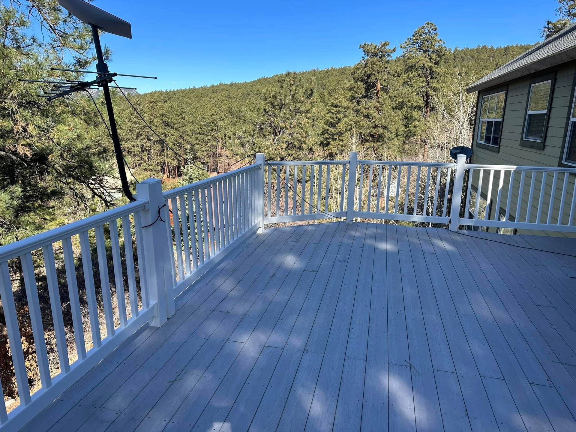 White wooden deck with railing overlooking a forest under a blue sky, next to a house.