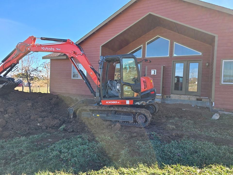 Red Kubota excavator digging near a red house with large windows.