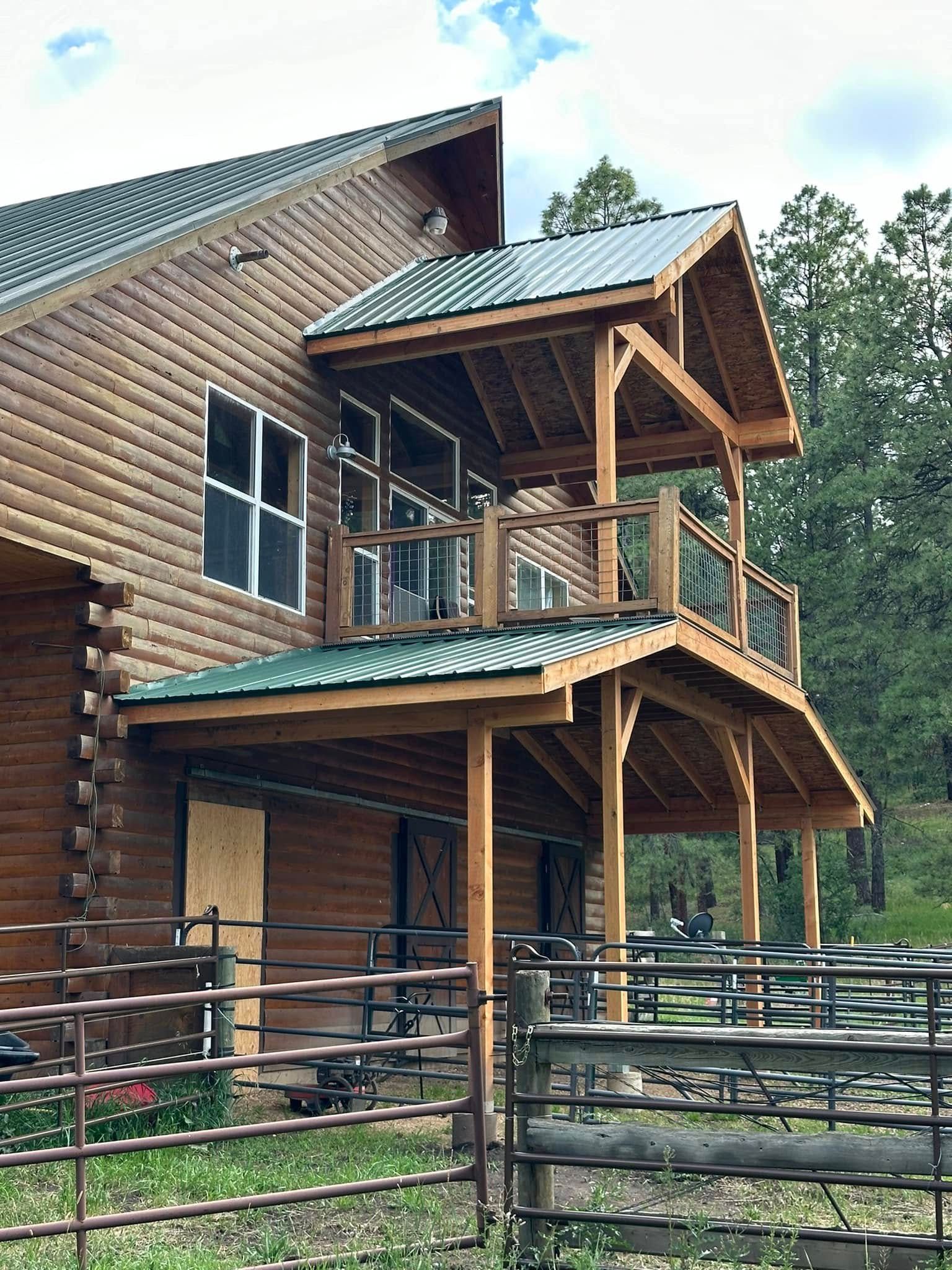 Log cabin with two-level wooden balcony and green metal roof, surrounded by a fence and trees.