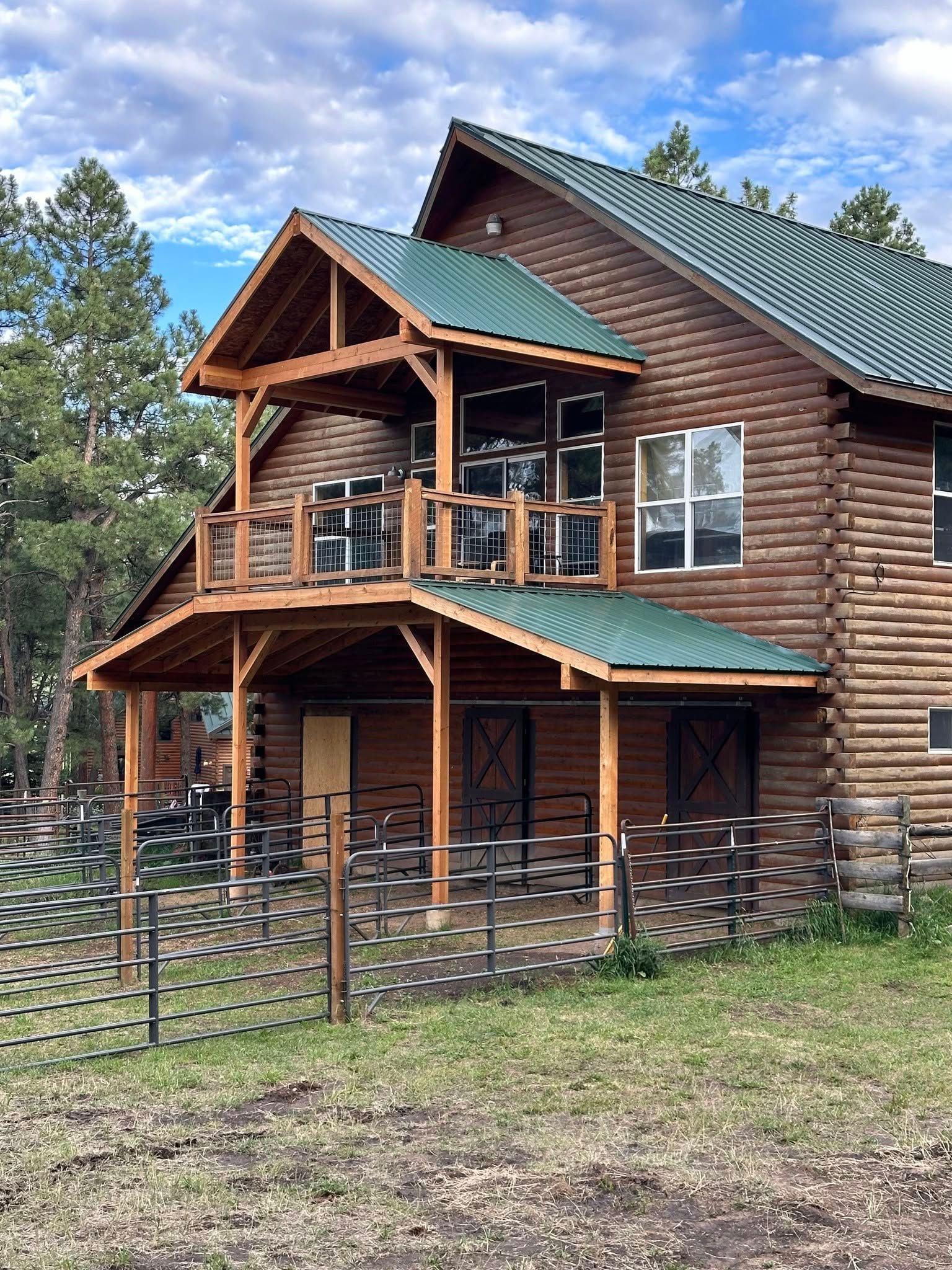 Log cabin with green roof, wooden balcony, set in a grassy area with a fence.