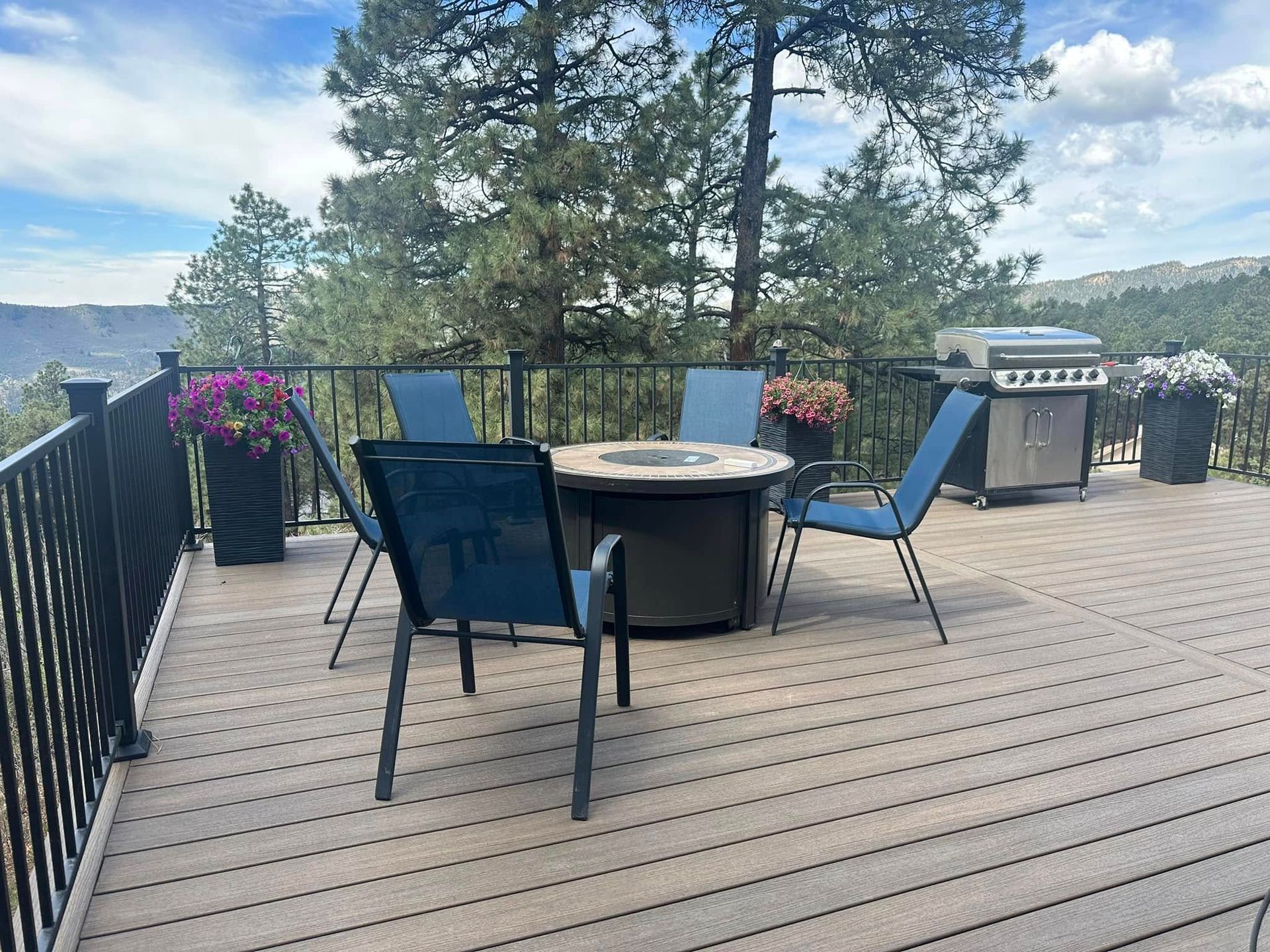 Deck with seating, fire pit, grill, and flower pots. Scenic view, blue chairs, and a sunny day.