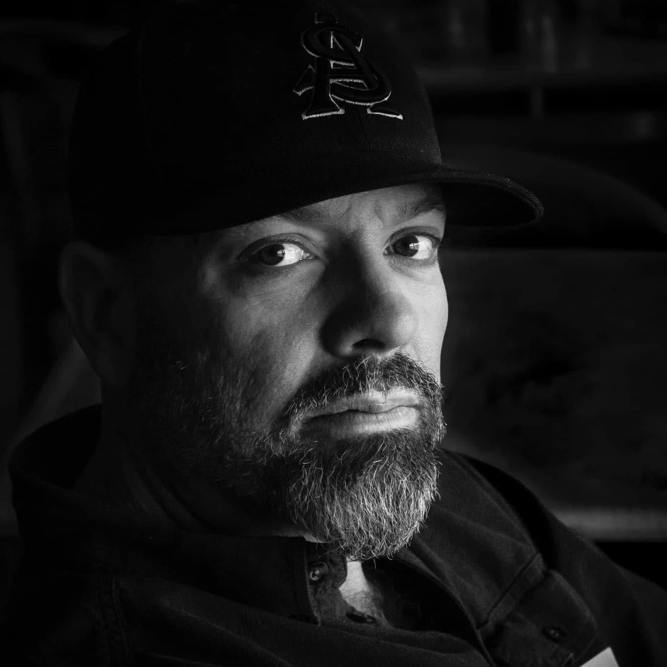 Man with a goatee wearing a baseball cap, looking directly at the camera. Black and white.