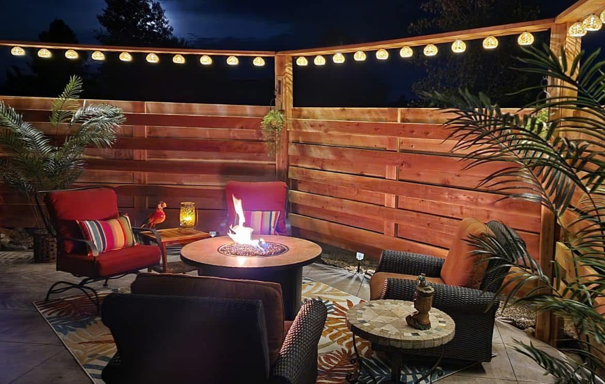 Nighttime outdoor patio with fire pit, seating, string lights, and wooden fence.