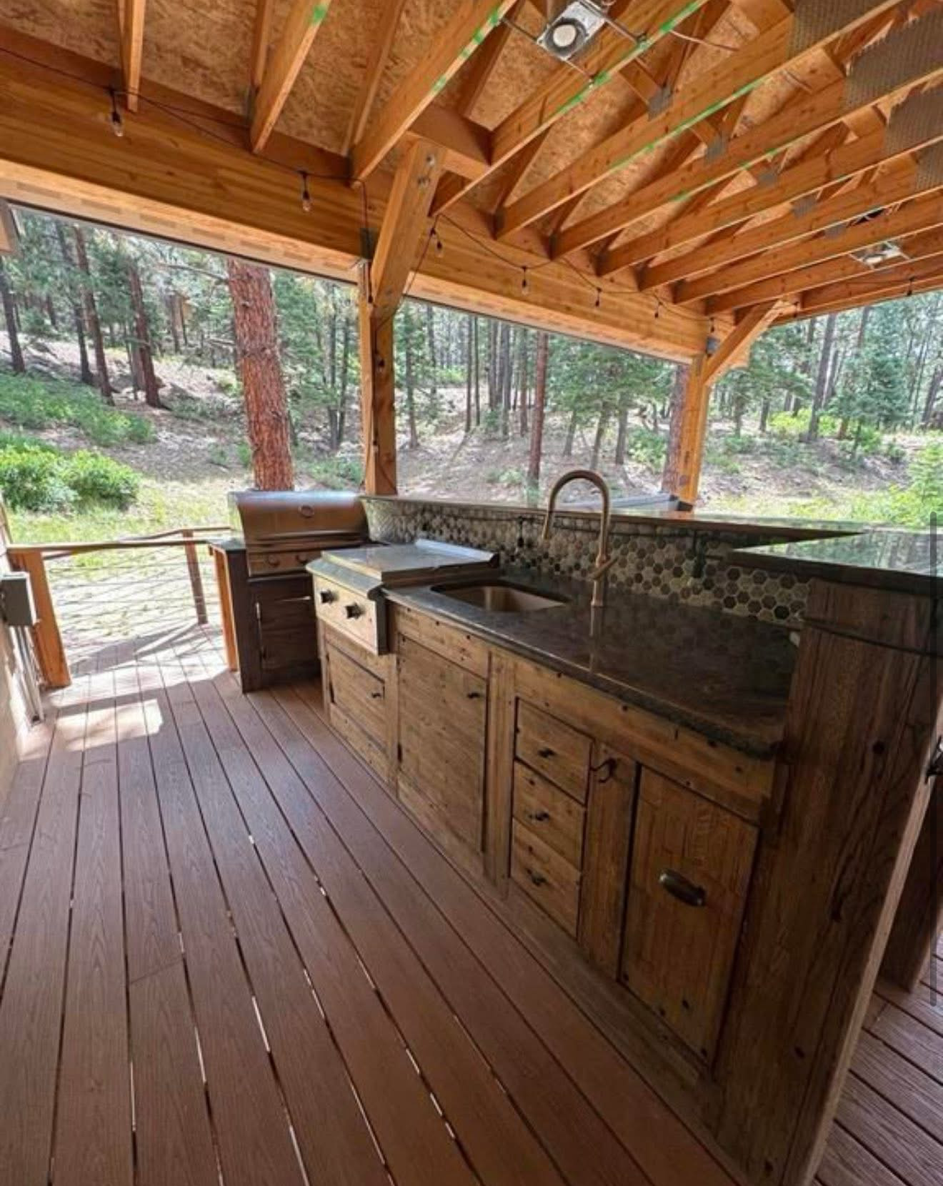 Outdoor kitchen with wooden cabinets, granite countertop, grill, and sink, under a wood-beamed overhang.