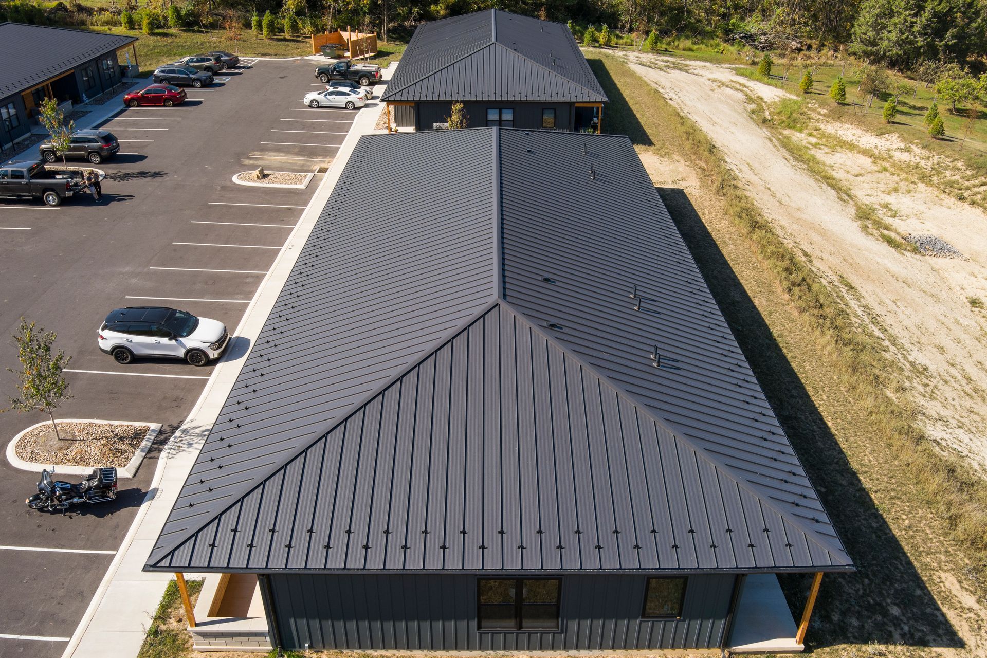 High-angle view of two dark metal-roofed buildings in a parking lot with several parked cars on a sunny day.