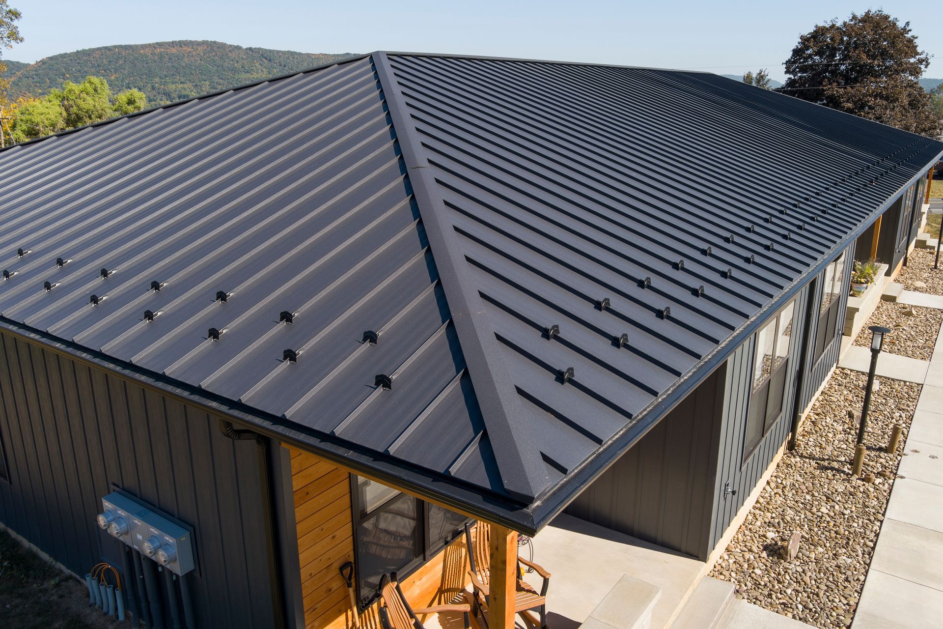 An aerial view of a dark metal roof on a modern home with a gravel side yard and distant hills under a clear blue sky.