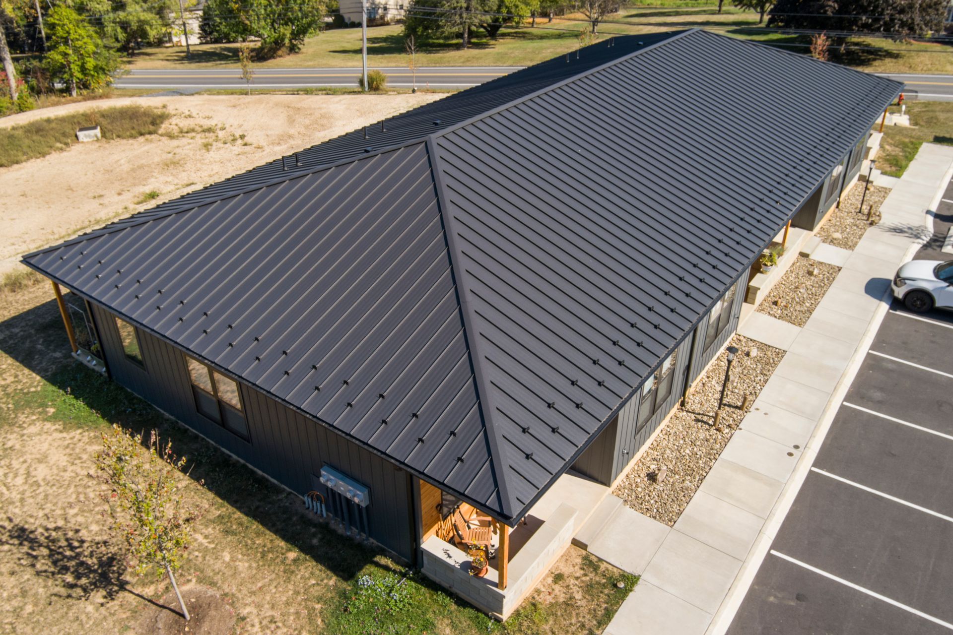 An aerial view of a single-story building with a dark metal roof, surrounded by landscaping, a sidewalk, and a parking lot.